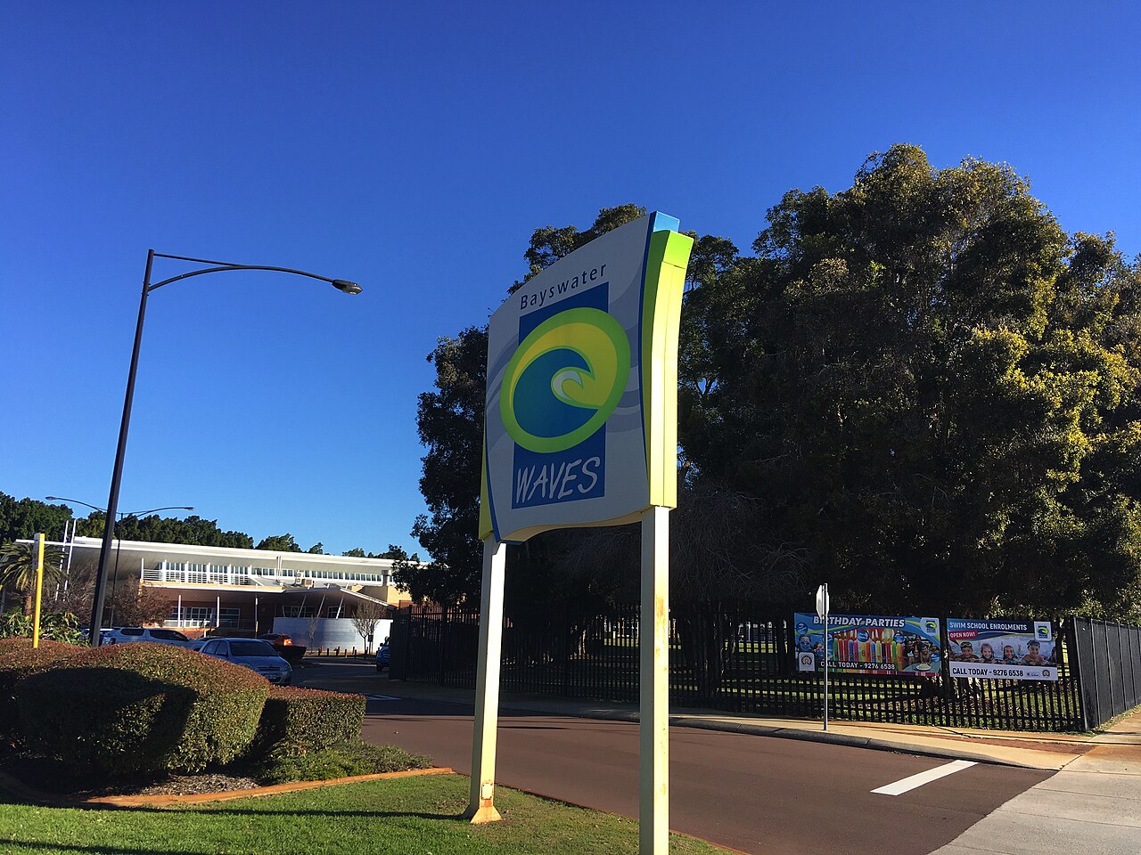 The Bayswater Waves sign beside the entry road, with the aquatic centre building and tall trees behind.