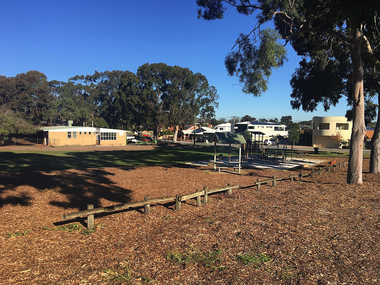 A park with mulch, low timber rails, a small brick building, a playground and houses beyond a stand of gum trees.