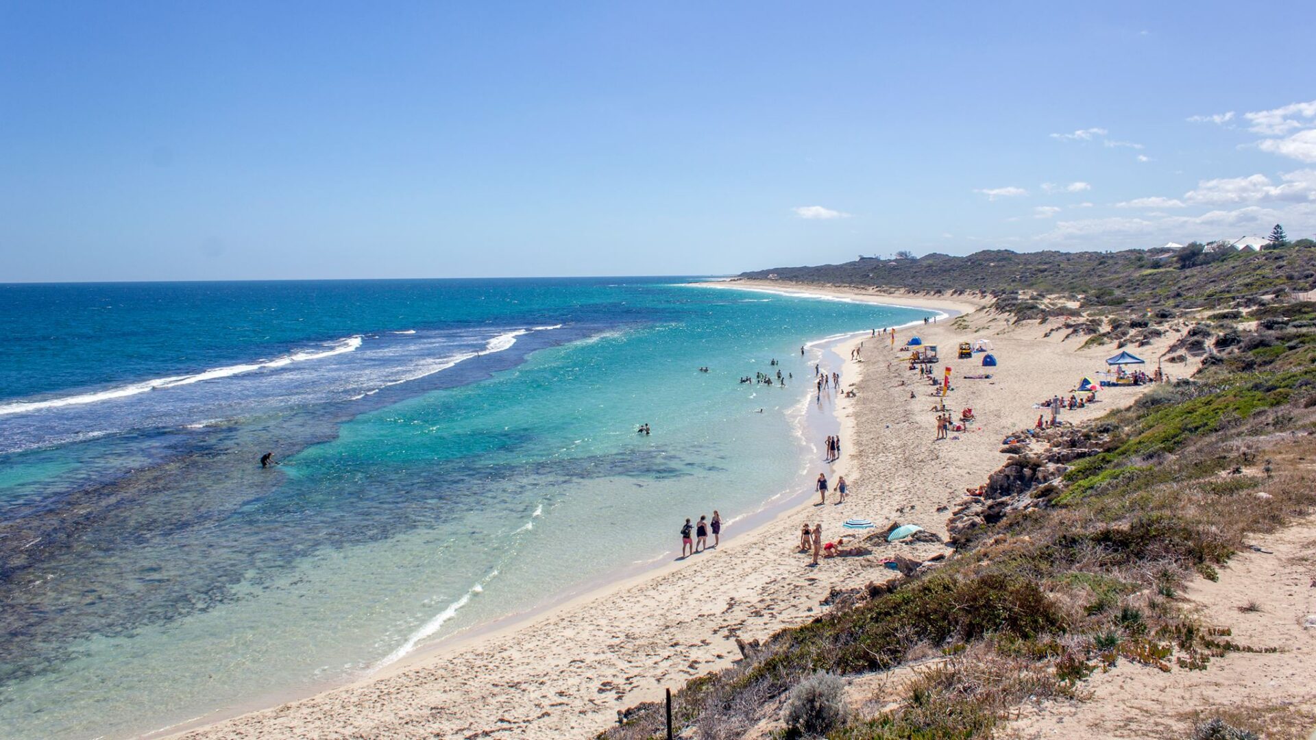 Looking along Yanchep Lagoon Beach from a dune, with turquoise water, a reef line, people on the sand and coastal scrub.